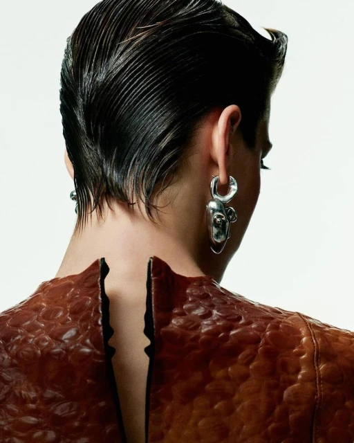 Woman's back view showcasing short, slicked-back hair, statement earrings, and a brown snakeskin-textured dress against a white backdrop.
