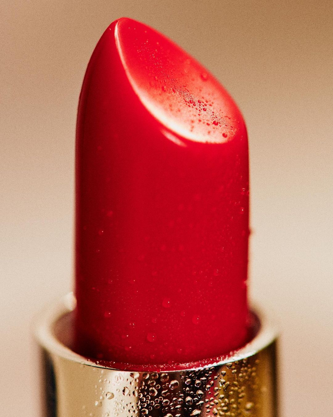 Macro close-up of a bright red lipstick bullet with condensation droplets on the metal base and body, against a warm neutral background.