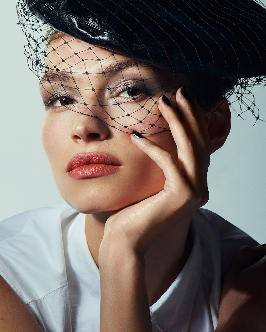 Close-up portrait of a woman with tan skin wearing a black leather hat and net veil, with glossy lips and black nail polish.
