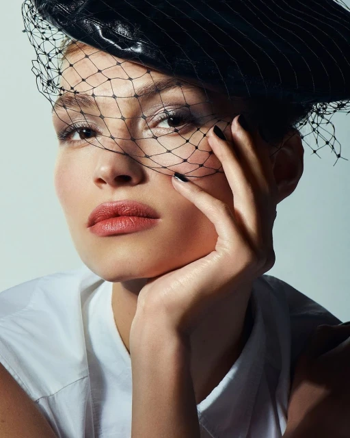 Close-up portrait of a woman with tan skin wearing a black leather hat and net veil, with glossy lips and black nail polish.