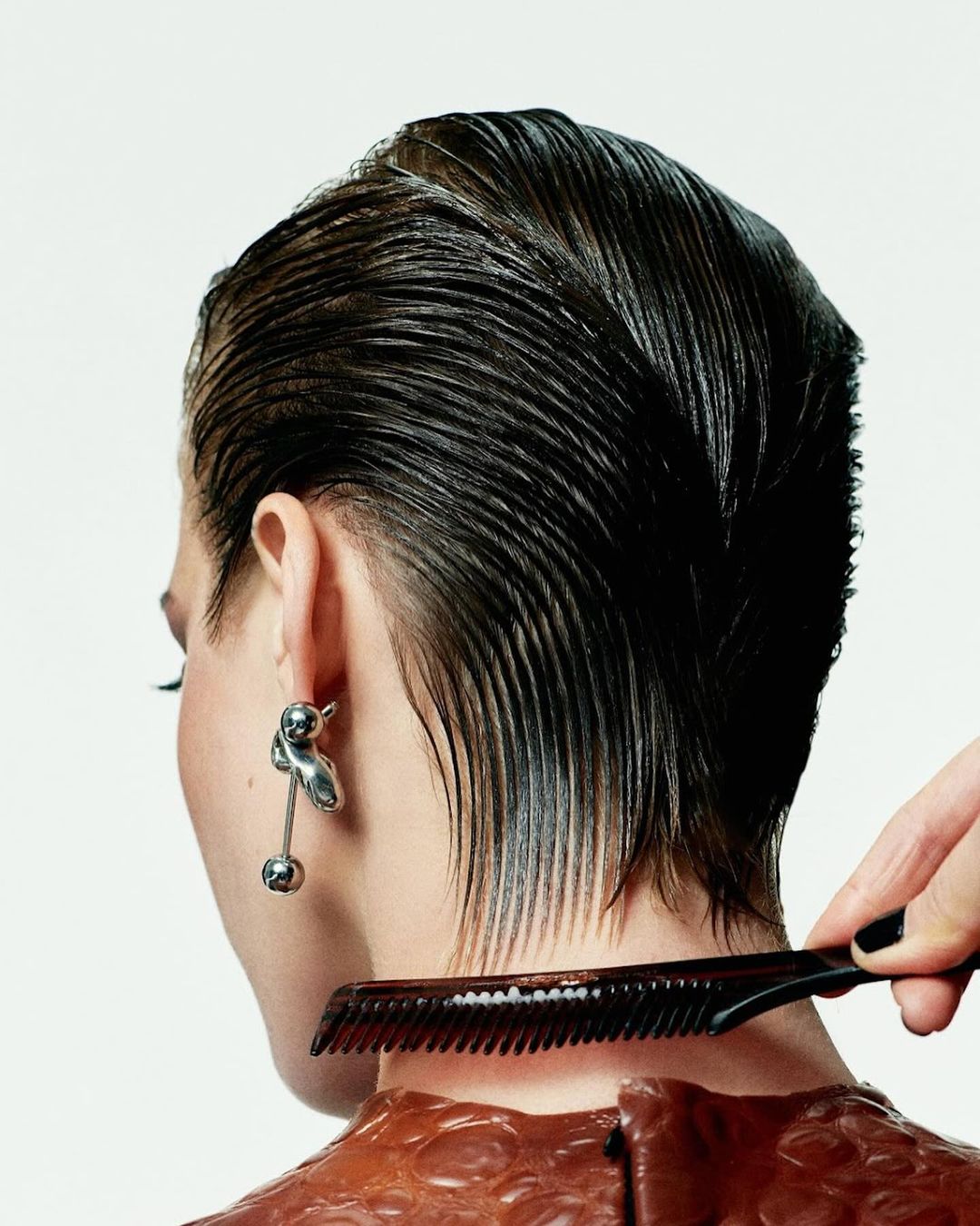 Back view of a person with dark, wet hair being styled with a comb against a white background in a beauty or fashion setting.