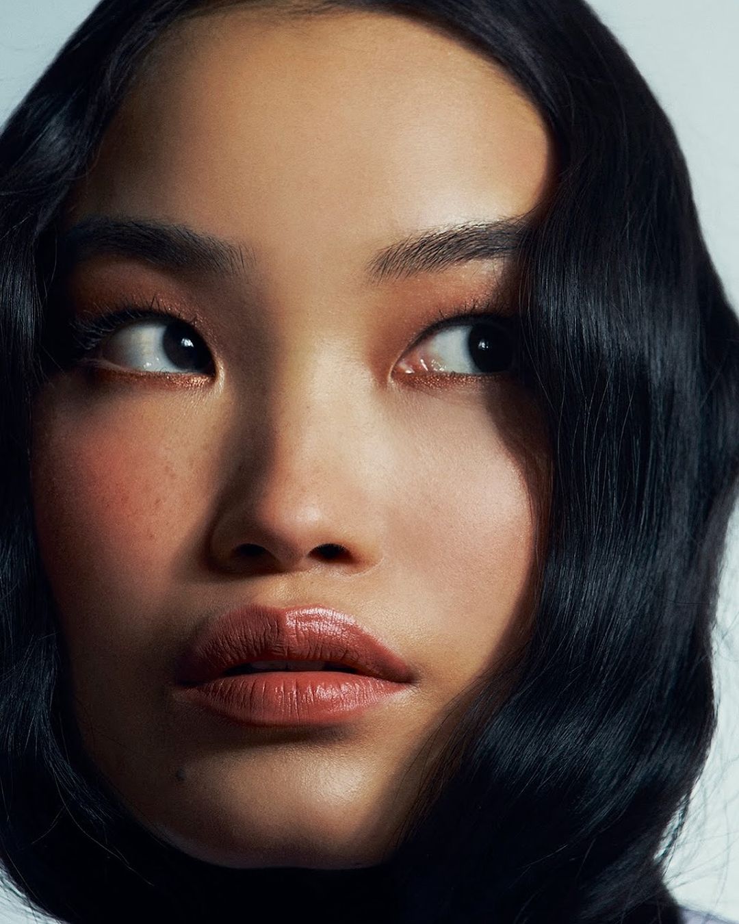 Close-up portrait of an Asian woman with dark wavy hair, glossy lips, and natural makeup looking off to the side against a white background.