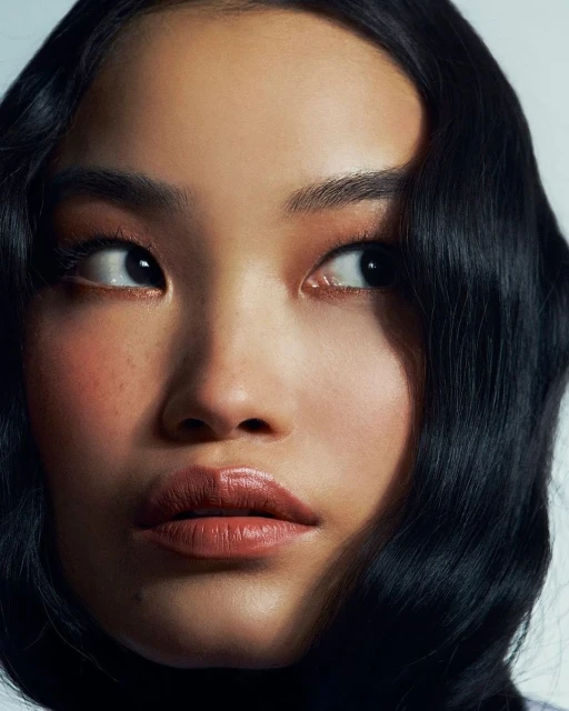 Close-up portrait of an Asian woman with dark wavy hair, glossy lips, and natural makeup looking off to the side against a white background.