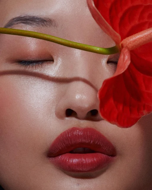 Close-up of a woman's face with closed eyes and red lipstick, partially obscured by a vibrant red Anthurium flower in soft studio lighting.