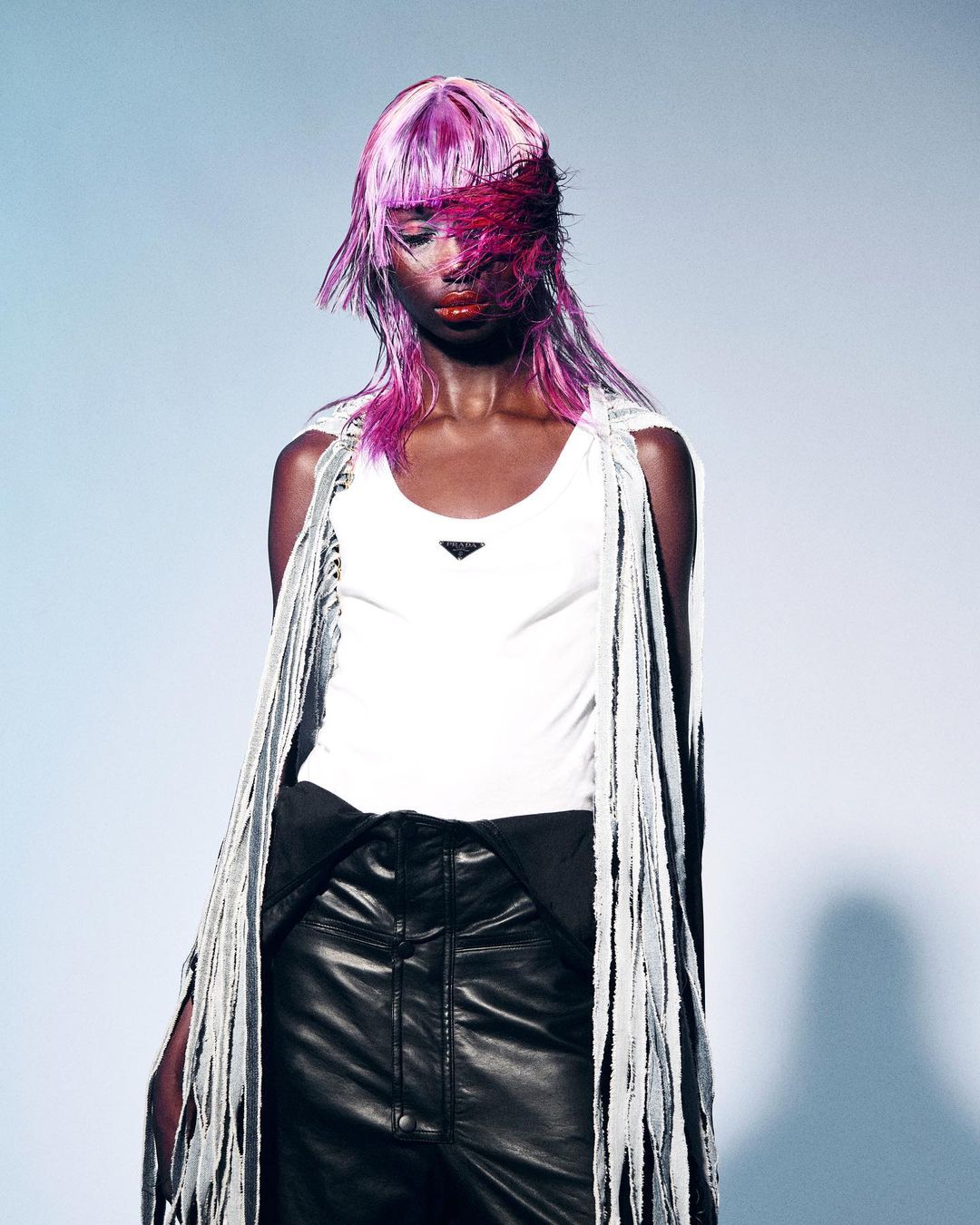 A fashion portrait of a black woman with pink hair wearing a white tank top and black leather pants in a light blue photo studio.