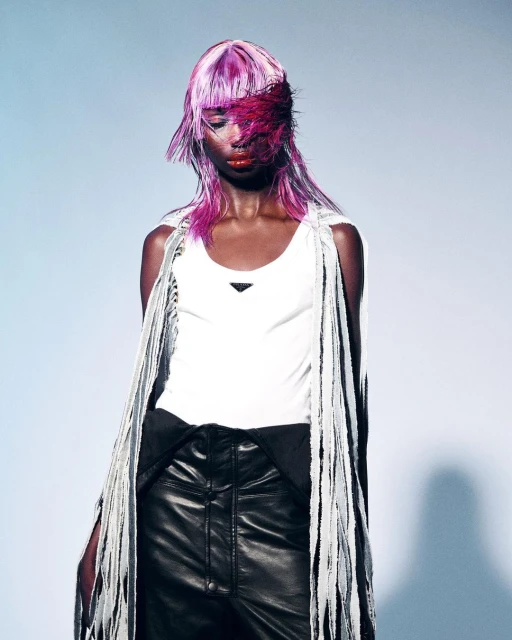 A fashion portrait of a black woman with pink hair wearing a white tank top and black leather pants in a light blue photo studio.