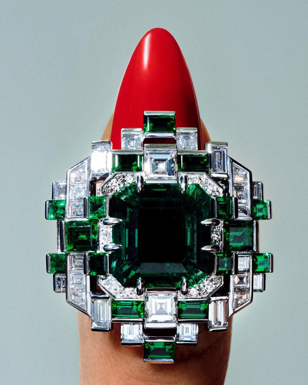 Macro shot of a finger wearing a geometric ring with an emerald surrounded by diamonds, set against a neutral gradient background.