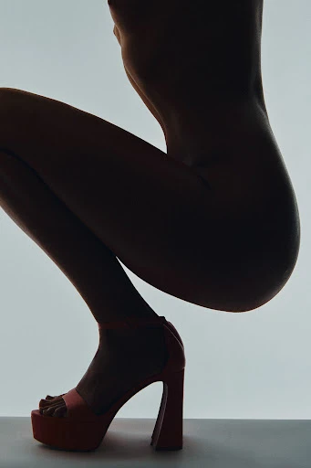 Silhouette of a nude adult female crouching in a studio against a bright background while wearing a pair of red platform heels.