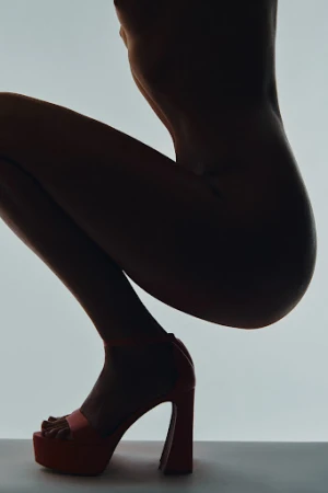 Silhouette of a nude adult female crouching in a studio against a bright background while wearing a pair of red platform heels.