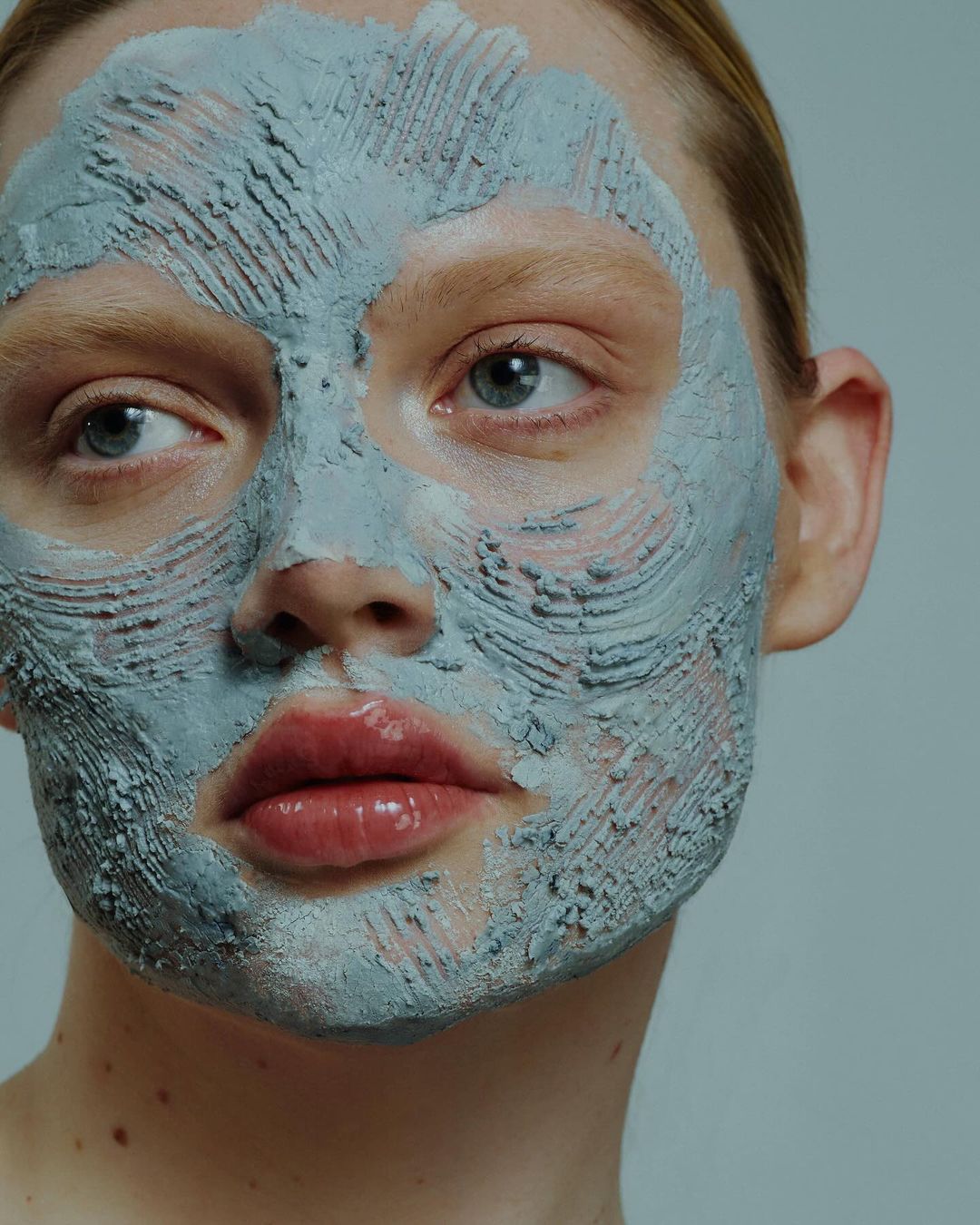 Close-up portrait of a blonde woman with a textured grey face mask. Neutral background. Focus on skincare and facial treatment.