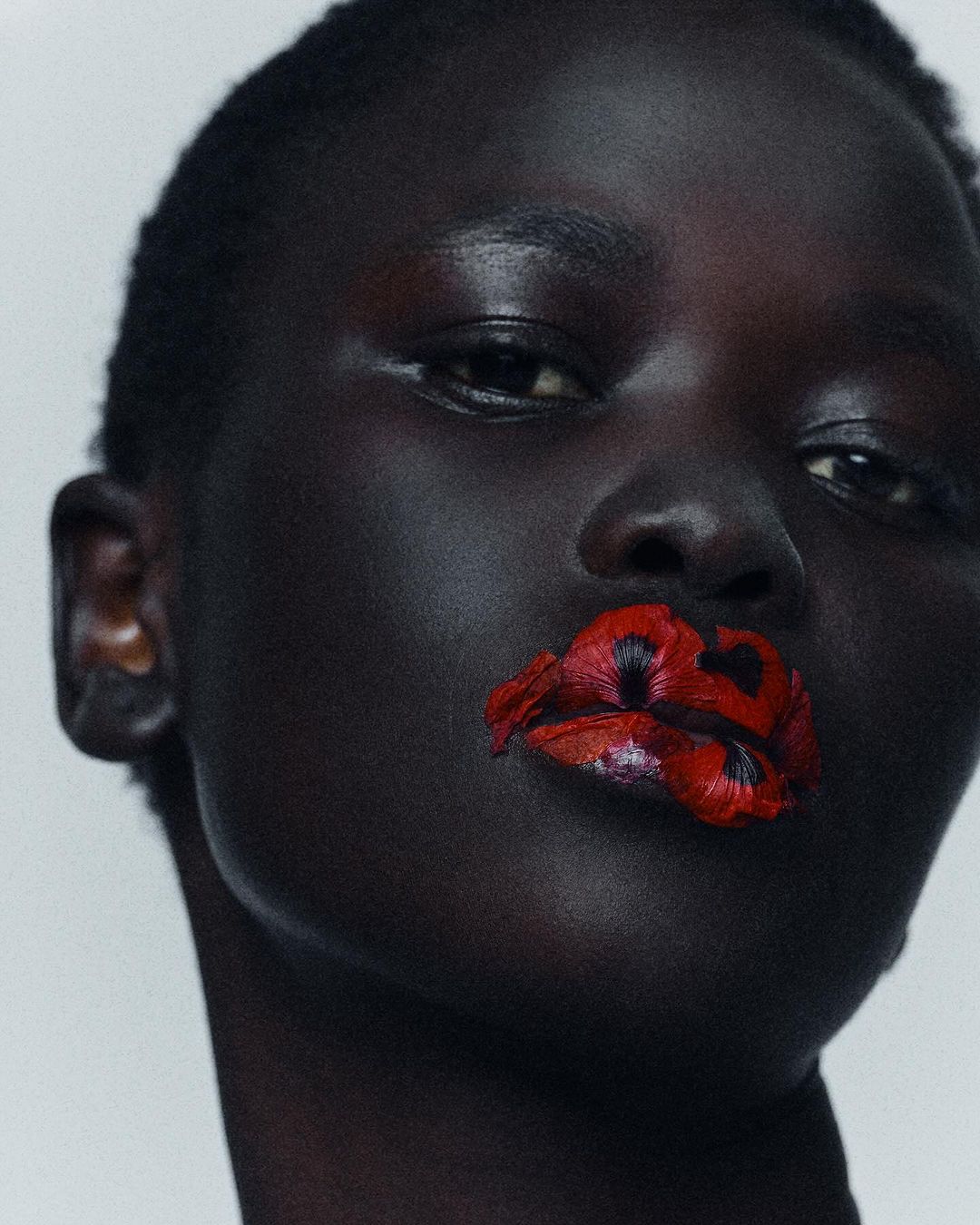 Close-up portrait of a Black woman with short hair wearing vibrant red floral petals adhered to her lips.