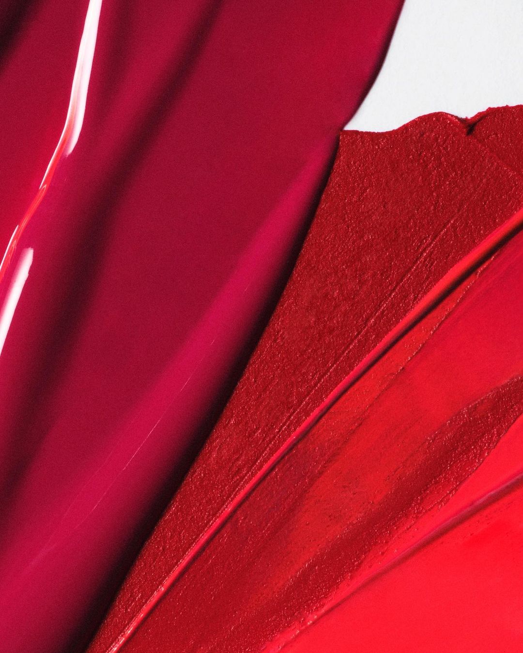 Close-up abstract view of red and burgundy lipstick swatches with glossy and matte textures on a white surface.