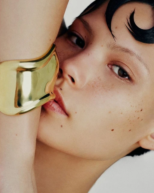 Close-up portrait of a young adult female model with freckles, glossy makeup, and slicked-back hair, wearing a wide gold cuff bracelet.