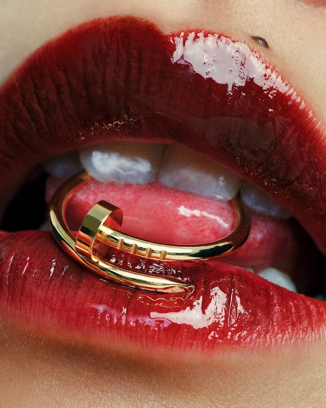 Close-up on person's mouth with glossy red lipstick and a gold nail-shaped ring between their teeth and tongue, fashion beauty shot.