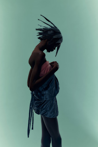 Fashion photo of a slender Black woman wearing avant-garde fashion with spiky hair, side view against a pale green background in studio lighting.