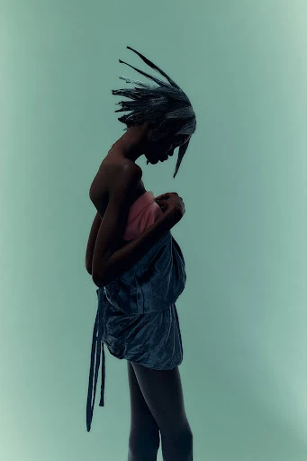 Fashion photo of a slender Black woman wearing avant-garde fashion with spiky hair, side view against a pale green background in studio lighting.