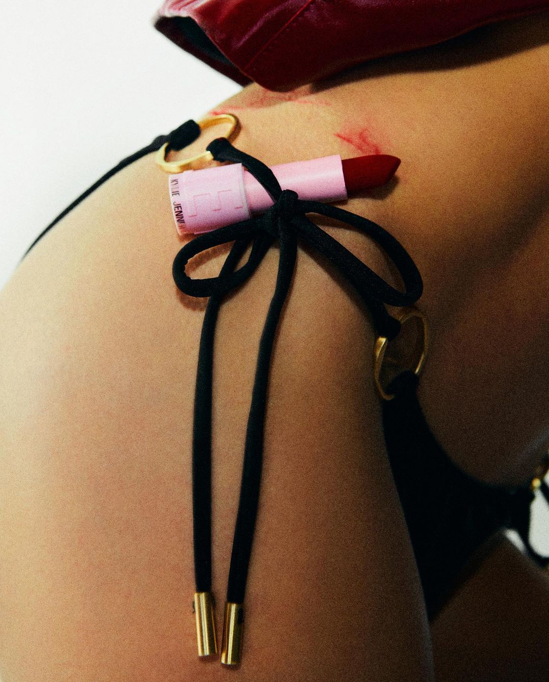 Close-up of a woman's body with a pink lipstick attached to her black bikini bottoms with gold hardware and red jacket.