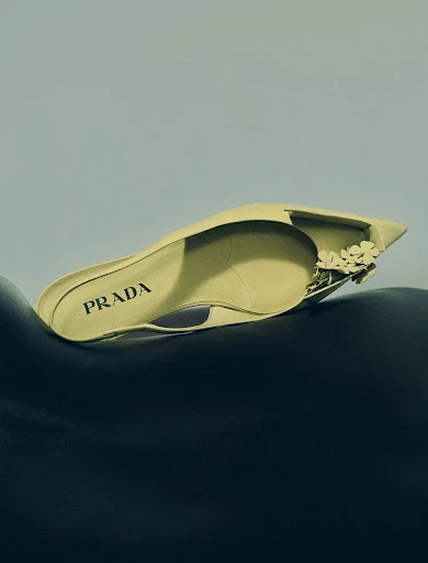 Elegant yellow Prada mule shoe with floral embellishments sits on a dark, curved surface against a soft blue background in a fashion still life.