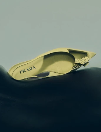 Elegant yellow Prada mule shoe with floral embellishments sits on a dark, curved surface against a soft blue background in a fashion still life.