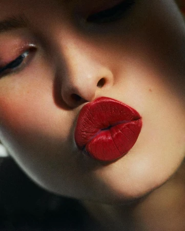 Close-up portrait of a woman puckering her vibrant red lips with subtle pink eyeshadow and a warm, grainy film texture.