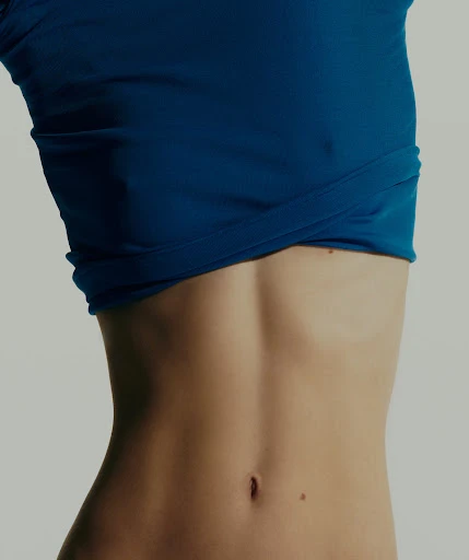 Torso of a woman in a blue top against a white background, close-up on her midriff and abdomen, showcasing fitness and fashion.