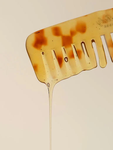 Close-up studio shot of a light-yellow-and-brown patterned hair comb with clear liquid dripping from it against a light background.