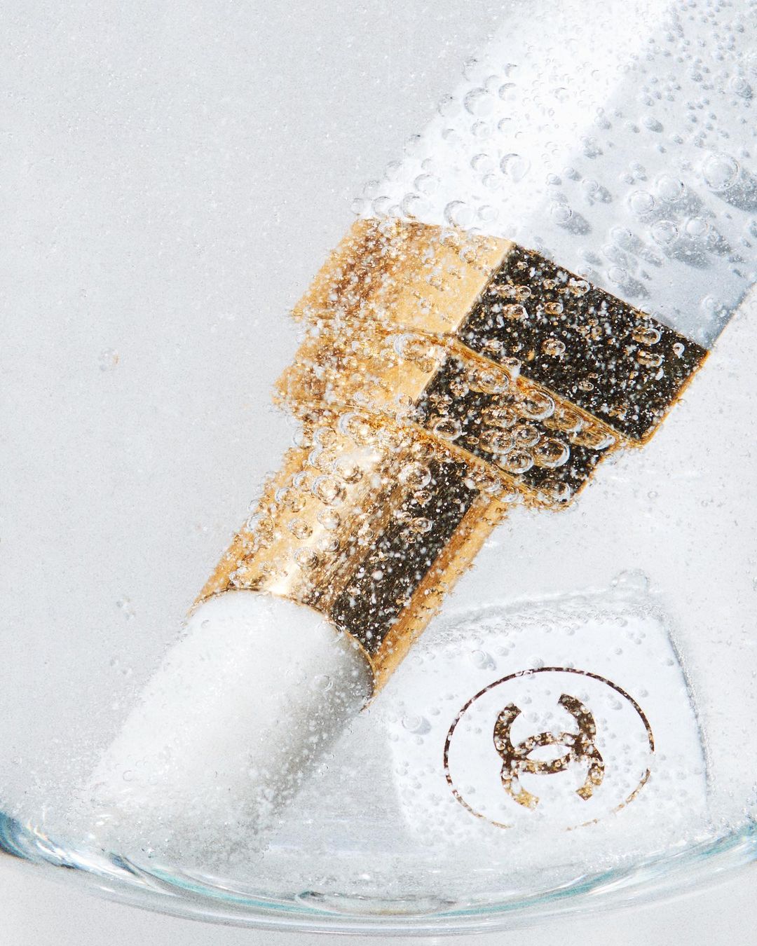 Close-up of a makeup brush with a gold handle and white bristles submerged in water with bubbles and the Chanel logo on a white container.