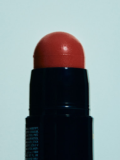 Close-up of a coral blush stick with a dark blue container against a pale bluish-white background, showcasing beauty product.