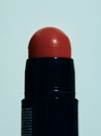 Close-up of a coral blush stick with a dark blue container against a pale bluish-white background, showcasing beauty product.