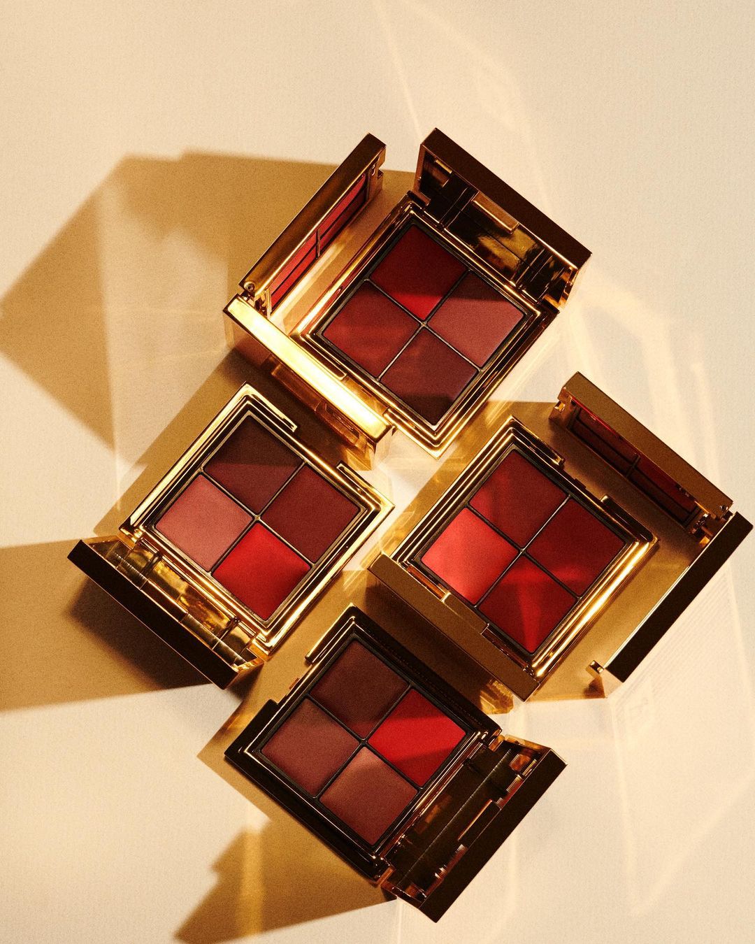 Four golden compacts with red-toned makeup sit on a white surface, casting dramatic shadows in a high-angle, studio product shot.