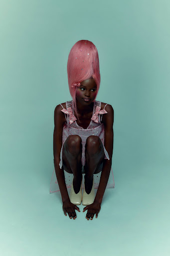 Young adult woman with dark skin and a pink wig sits on the floor in a light pink dress and cream shoes against an aqua-colored backdrop.