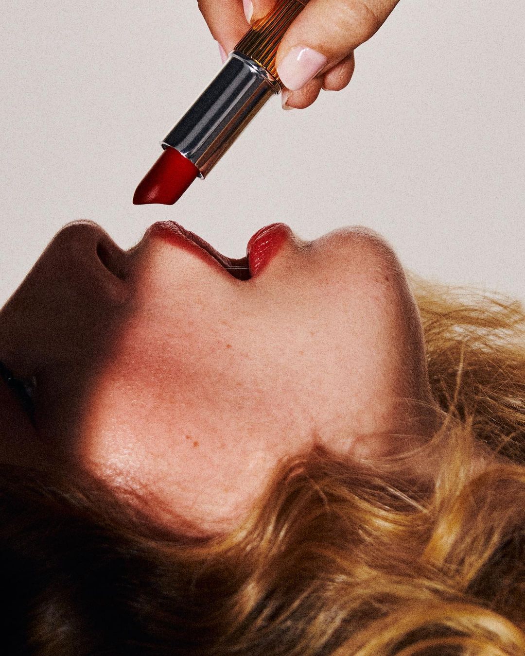 Close-up of a hand holding red lipstick over a woman's face, focusing on the lips and the product application in a beauty context.