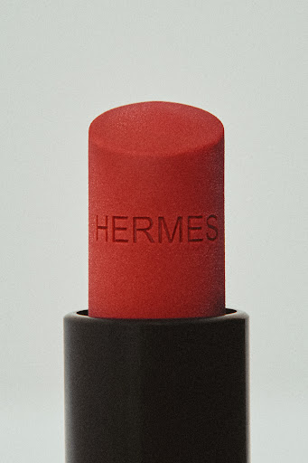 Close-up of a red lipstick tube with the brand name HERMES embossed on it against a plain light gray background, beauty shot.