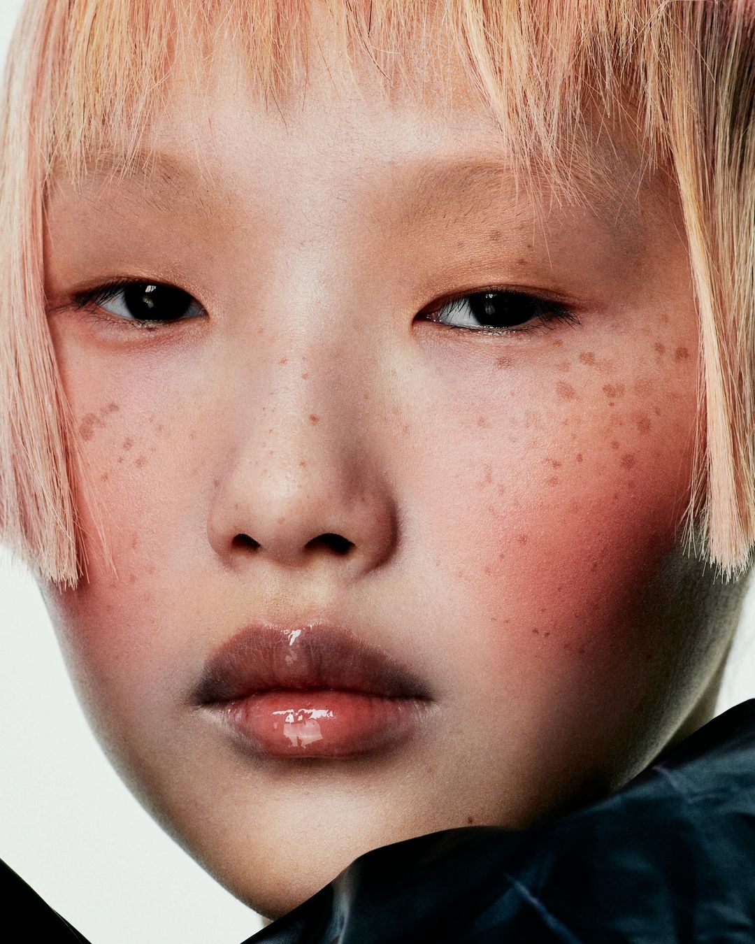 Close-up portrait of a young Asian female with light pink hair, freckles, glossy lips, and wearing a black textured garment against a white background.