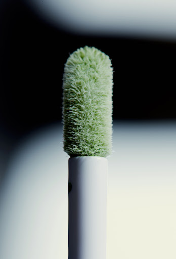 Close-up of a light-green, fuzzy makeup applicator with a white handle, set against a blurred background of black and light gray.