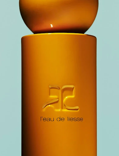 Close-up studio shot of a yellow and orange perfume bottle with the brand logo and 'l'eau de liesse' text on a light blue backdrop.