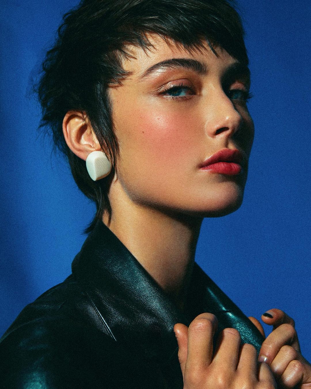 Close-up portrait of a woman with short dark hair, blue eyes, and glossy red lips, wearing a black jacket and white earrings against a blue backdrop.