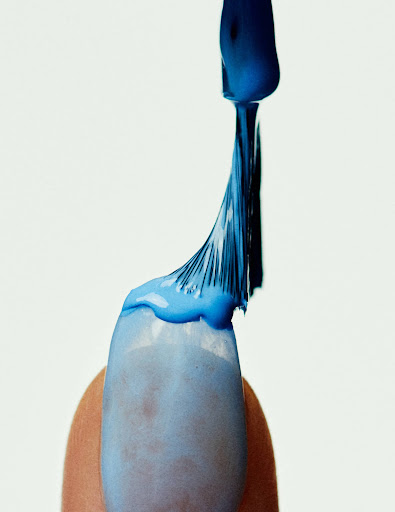 Close-up of blue nail polish being applied to a fingernail against a white background, emphasizing the texture and color.
