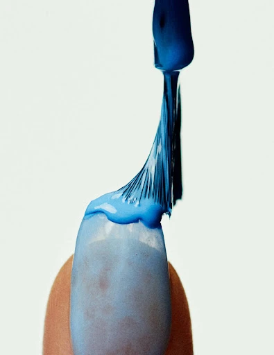 Close-up of blue nail polish being applied to a fingernail against a white background, emphasizing the texture and color.