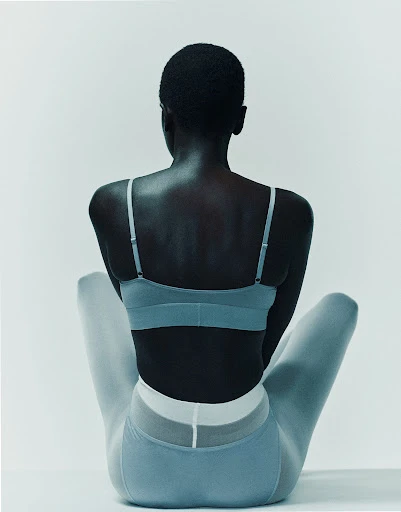 Back view of a Black female model in a light blue sports bra and leggings against a white backdrop in a fashion studio.