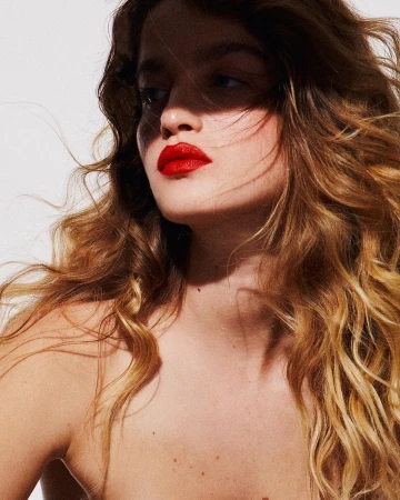 Close-up portrait of a woman with long wavy blonde hair and vibrant red lipstick under direct shadow lighting against a light background.