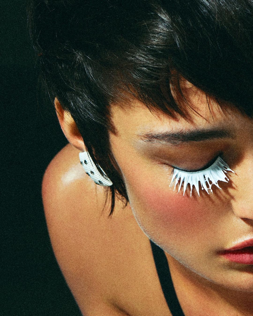 Close-up beauty shot of a model with short dark hair and white eyelashes, wearing a spotted earring, against a dark backdrop.