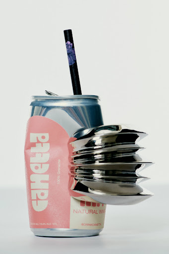 A crushed can with a straw and a silver metal object sits on a white background in a studio lighting.