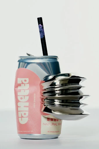 A crushed can with a straw and a silver metal object sits on a white background in a studio lighting.