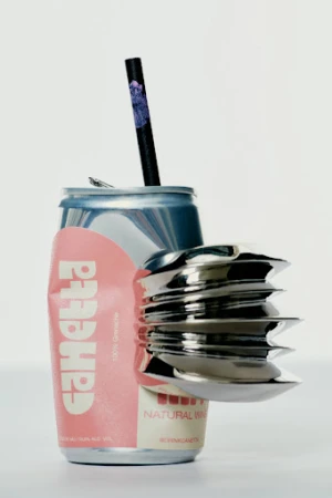 A crushed can with a straw and a silver metal object sits on a white background in a studio lighting.