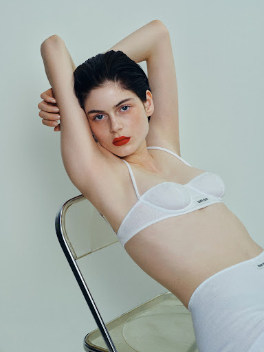 Woman with red lipstick poses in white lingerie while leaning back on a clear plastic chair against a light-grey studio backdrop.