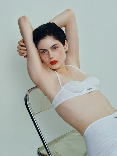 Woman with red lipstick poses in white lingerie while leaning back on a clear plastic chair against a light-grey studio backdrop.