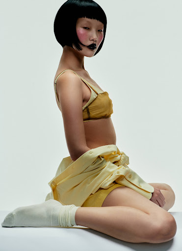 Asian woman with short black bob and yellow outfit poses against white backdrop in fashion shot with bright, neutral lighting and cool tones.