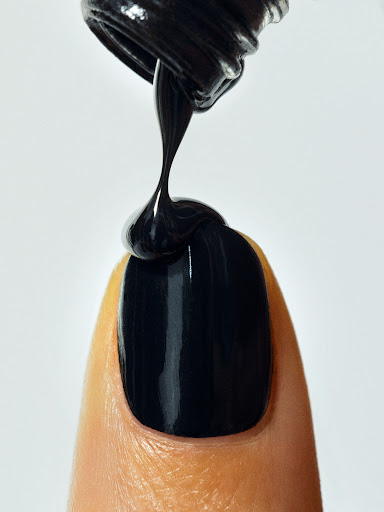 Close-up of black nail polish being applied to a fingernail, set against a clean white background with focus on the polish application.