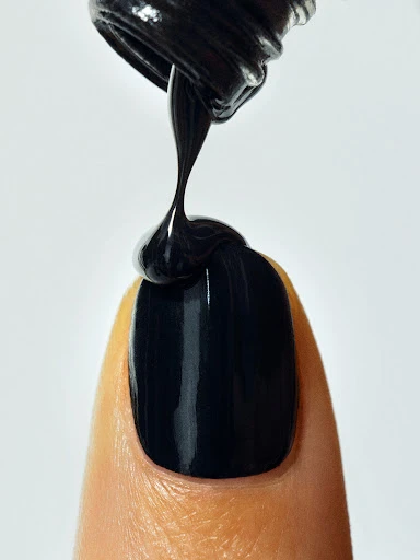 Close-up of black nail polish being applied to a fingernail, set against a clean white background with focus on the polish application.